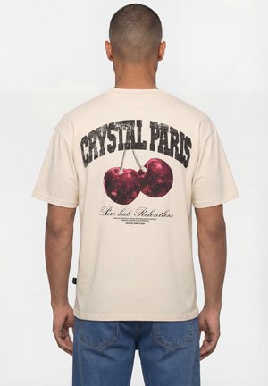 Uomo che indossa una maglietta beige con grandi ciliegie rosse e il testo "CRYSTAL PARIS Pure but Relentless" sulla schiena, abbinata a jeans blu.