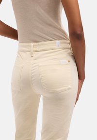 Person iført beige tætsiddende bukser og en ærmeløs top, vist fra taljen og ned med hænderne hvilende ved siden.