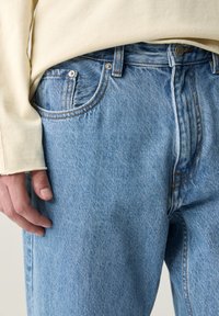 Gros plan sur une personne portant un jean en denim bleu clair et un haut à manches longues crème, avec la main gauche reposant le long du corps.