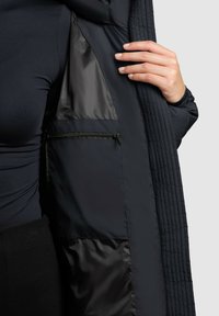 Prošívaná černá bunda s lesklým interiérem, na kterém je vidět boční kapsa se zipovým uzávěrem. Detail ruky držící bundu.