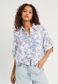 Chemise blanche à boutons avec un motif cachemire bleu, manches courtes retroussées et coupe décontractée, associée à un jean en denim bleu clair.