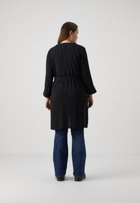 Robe noire à manches longues avec une ceinture à nouer, présentant une coupe droite et une légère fente au dos, associée à un jean bleu évasé et des chaussures noires.
