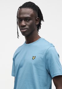 Light blue cotton t-shirt with a crew neck and short sleeves. Features a small black and yellow bird logo on the left chest area.