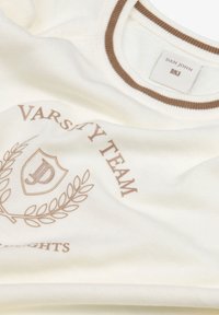 Sudadera de color crema con un cuello acanalado marrón. En la parte delantera muestra "VARSITY TEAM" y un emblema con detalles de laurel.