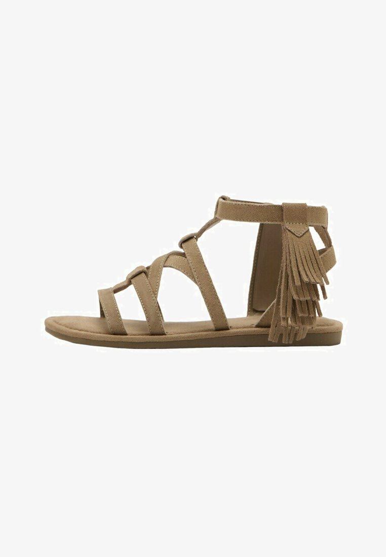 Sandale gladiateur plate en daim beige avec bride à la cheville et multiples franges décoratives à l'arrière.