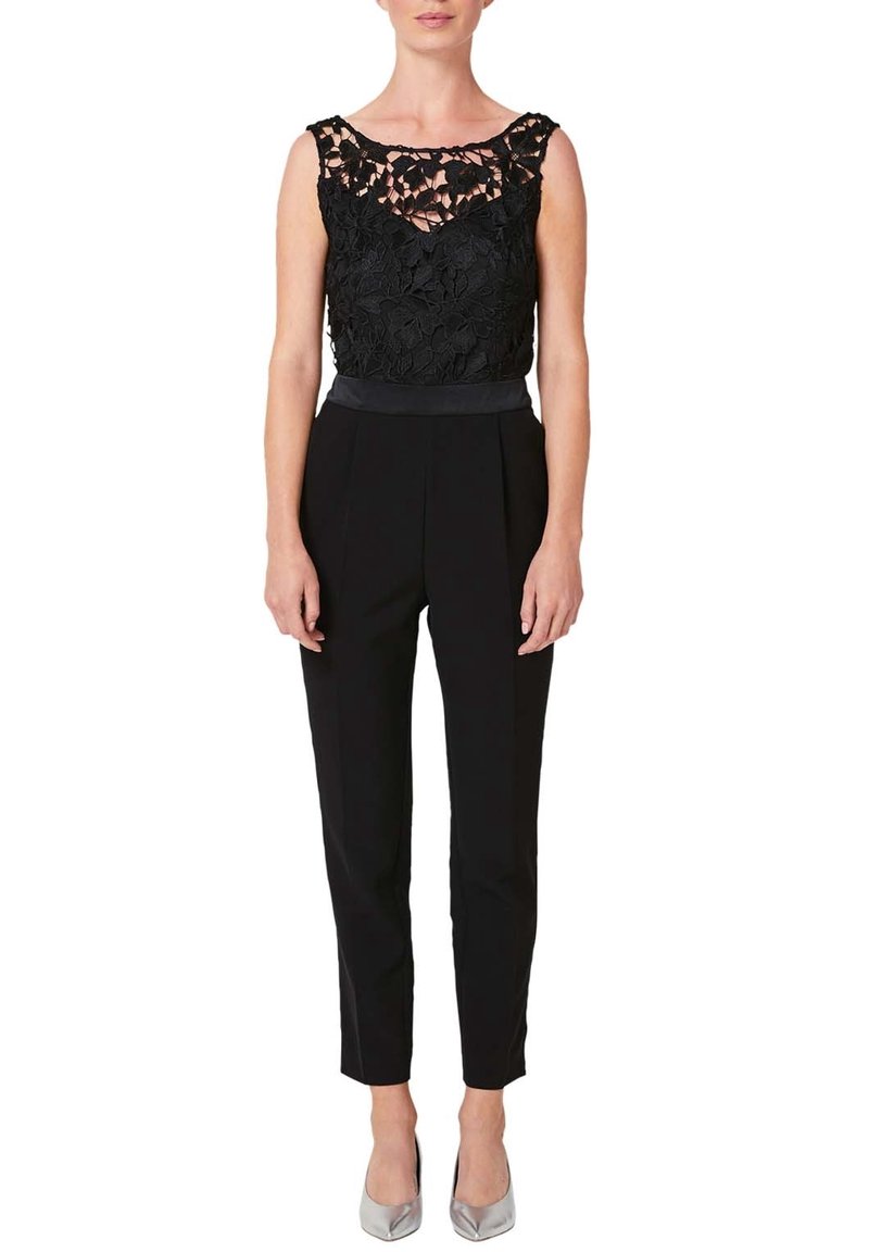 Femme portant un haut en dentelle noire sans manches associé à un pantalon noir taille haute fuselé et des talons pointus argentés sur fond blanc.