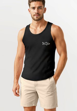 Schwarzes Tanktop mit einem weißen Haifischgrafik auf der linken Brust, aus leichtem Stoff; kombiniert mit beigen Shorts, die eine lockere Passform zeigen.