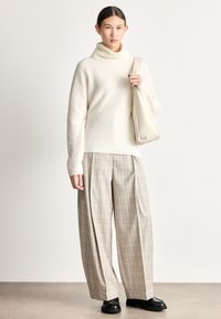 Camisola creme de gola alta com textura canelada, combinada com calças largas de padrão às quadrículas em bege e castanho claro. Sapatos pretos e uma mala bege.