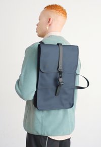 Marineblå rygsæk med en topklap, central rem og justerbare sideremme. Lavet af glat materiale med minimalistisk design.