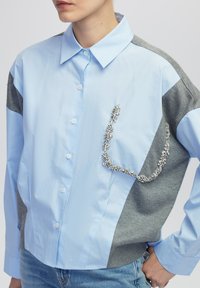 Camisa azul claro y gris de botones, con cuello, un bolsillo frontal adornado con detalles de cristal y mangas enrolladas de forma casual.
