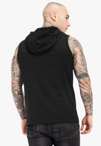 Melnā bezpiedurkņu hoodie ar kengura kabatu un teksturētu audumu. Tam ir mīksta kapuce un uz rokām ir redzami izteikti tetovējumi.