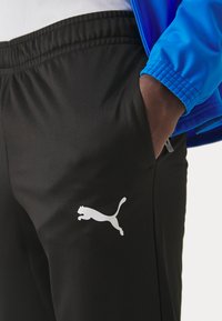 Zwarte sportbroek met een gladde textuur, met een witte Puma-logo aan de zijkant. Een blauwe jas is gedeeltelijk zichtbaar aan de bovenkant.