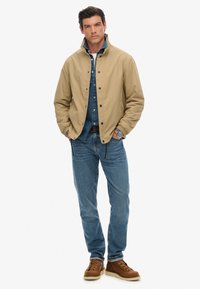 Lichtbruine jas met drukknoopjes over een donkerdenim shirt, gecombineerd met blauwe jeans en bruine veterschoenen. Model staat rechtop.