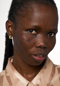 Närbild av en person med mörk hud, iförd en ljusbeige satinskjorta och guldörhängen i hoop-stil. Huden visar textur och subtila fläckar.