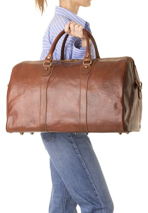 Persona con una camicia a righe blu e jeans che porta una grande borsa da viaggio in pelle marrone tenendola per le maniglie.