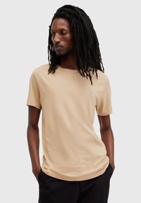 T-shirt beige a maniche corte in cotone, con scollatura rotonda e un piccolo logo sul petto. Il modello posa con le mani in tasca.