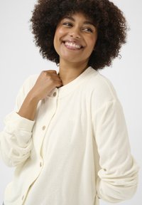 Cardigan color crema realizzato in materiale morbido, con chiusura a bottoni, scollatura rotonda e maniche lunghe leggermente a palloncino con una finitura testurizzata.