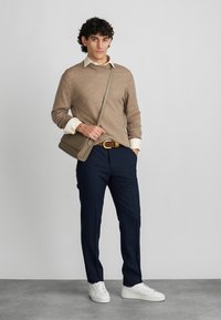 Bēša adīta džemperis, zem tā balts kreklis ar apkakli, tumši zili bikses, brūns jostas, un pelēka rokas soma. Balti sporta apavi noslēdz apģērbu.