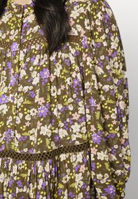 Vestido floral em tecido castanho com flores roxas, rosas e amarelas. Apresenta mangas bufantes e uma cintura texturizada com costura detalhada.