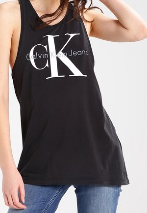 Osoba w czarnym bezrękawniku Calvin Klein Jeans z dużym białym logo "CK", w niebieskich dżinsach na jasnym tle.