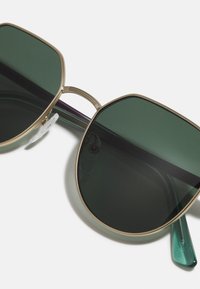 Ochelari de soare cu rame aurii, lentile mari de culoare verde închis și suporturi pentru nas transparente. Temele sunt accentuate cu o nuanță translucidă de teal.