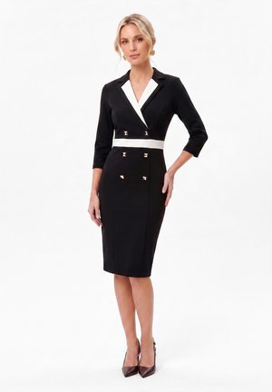 Femme portant une robe noire longueur genou avec un col blanc et un détail à la taille, des boutons dorés, des manches trois quarts et des talons pointus marron.
