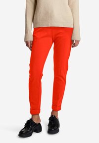 Rote, taillierte Hosen aus einem dehnbaren Material, mit einem umgeschlagenen Saum. Kombiniert mit schwarzen, klobigen Schuhen und einem hellbeigen Pullover.