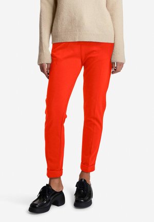 Pantalons rouges fuselés en tissu extensible, avec un ourlet à revers. Associés à des chaussures noires à semelle épaisse et un pull léger beige.
