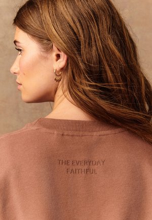 Frau mit langen braunen Haaren, die einen braunen Pullover mit dem eingeprägten Text „THE EVERYDAY FAITHFUL“ auf dem Rücken und goldene Creolen trägt.