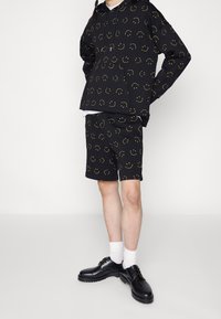 Sweat-shirt à capuche noir et shorts avec imprimé de visages souriants jaunes. Fabriqué en tissu doux, il possède une poche kangourou et une coupe décontractée. Chaussures noires.