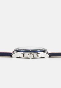 Reloj con correa de textil azul que presenta una caja de acero inoxidable, bisel azul, herraje plateado y una esfera blanca redonda con marcadores de hora.