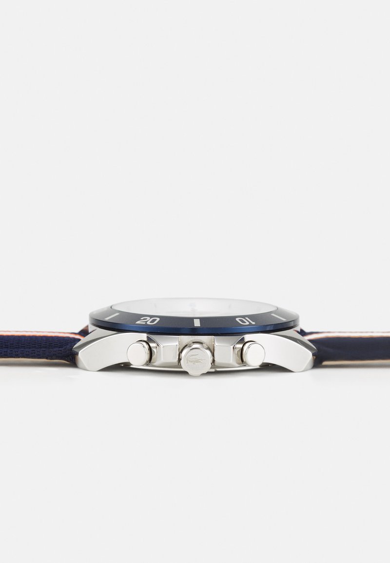 Reloj con correa de textil azul que presenta una caja de acero inoxidable, bisel azul, herraje plateado y una esfera blanca redonda con marcadores de hora.