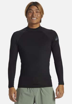 Rash guard noir à manches longues en matériau extensible, avec un col rond et des coutures plates ; il présente un petit logo gris sur l'épaule gauche.