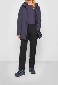 Vattenavvisande mörklila parkas med huva, lager på lager över en åtsittande lila topp, kombinerat med svarta byxor och blå vandringskängor.