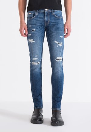 Faded blue denim jeans with distressed rips, slim fit design, five-pocket styling, and a button fly, paired with black ankle boots.