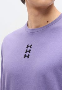 Άτομο που φοράει λιλά πουκάμισο με τρία μαύρα λογότυπα Under Armour τοποθετημένα κατακόρυφα στο στήθος και εμφανή κύστη στον λαιμό.