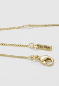 Dwa łańcuszki w kolorze złota o różnych stylach ogniw, z zapięciem typu karabińczyk i małą zawieszką z wygrawerowanym napisem "PILGRIM" na białym tle.