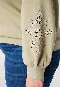 Lichtgroene sweatshirt met pofmouw en verfijnde witte bloemrijke borduursels, met uitsnijdingen. Draagt een blauwe spijkerbroek.