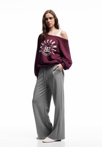 Mörkröd off-shoulder sweatshirt med grafiken "California 82"; grå vida träningsbyxor; enkla vita skor; slät textur.