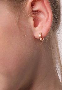 Gouden oorbel in de vorm van een ring met een textuurdesign en kleine versieringen, getoond gedragen aan het oor, waarbij de ronde vorm en details worden benadrukt.