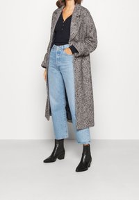 Grijze lang hondentandjas over een zwart topje, gecombineerd met lichtblauwe hoge taille jeans en zwarte enkellaarzen.