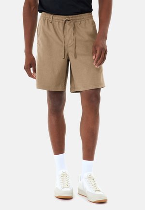 Uomo che indossa pantaloncini beige con coulisse, calzini bianchi e scarpe da ginnastica bianche, in piedi contro uno sfondo chiaro e uniforme.