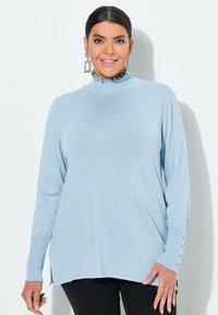 Hellblaues Langarmoberteil mit hohem Rüschenausschnitt, gerippten Bündchen mit Knöpfen und Seitenschlitzen für mehr Bewegungsfreiheit.