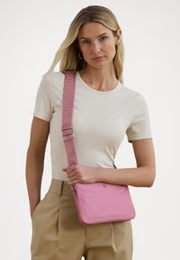 Pink crossbody bag with smooth texture, adjustable strap featuring branding, and a zip closure. Compact rectangular shape.