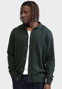 Un jeune homme à la peau foncée et aux dreadlocks teintées porte un cardigan vert foncé boutonné sur une chemise blanche, regardant vers la gauche avec les mains dans les poches.