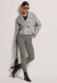 Graue, doppelreihige, kurze Jacke, schwarzes strukturiertes Oberteil, schwarz-weiße gemusterte Hose, schwarze Stiefeletten und kleine Handtasche.