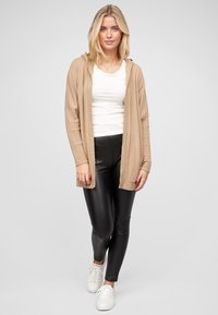 Beige gebreide cardigan met capuchon, geribbelde mouwen, gecombineerd met zwarte leren leggings en witte sneakers, voor een neutrale achtergrond.