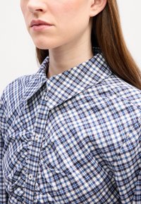 Camicia a quadri blu e bianchi con un ampio colletto, caratterizzata da pieghe verticali sul davanti e bottoni bianchi su un tessuto leggero.