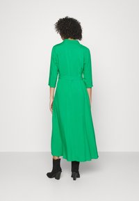 Grünes, langärmliges Kleid mit eng anliegendem Taillenbereich und A-Linien-Rock, aus einem glatten Stoff gefertigt. Getragen über schwarzen Knöchelstiefeln.