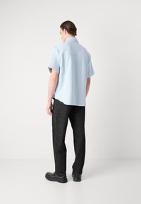 Camicia azzurra chiara a maniche corte con una vestibilità rilassata, abbinata a pantaloni in denim neri e scarpe nere, vista dal retro.